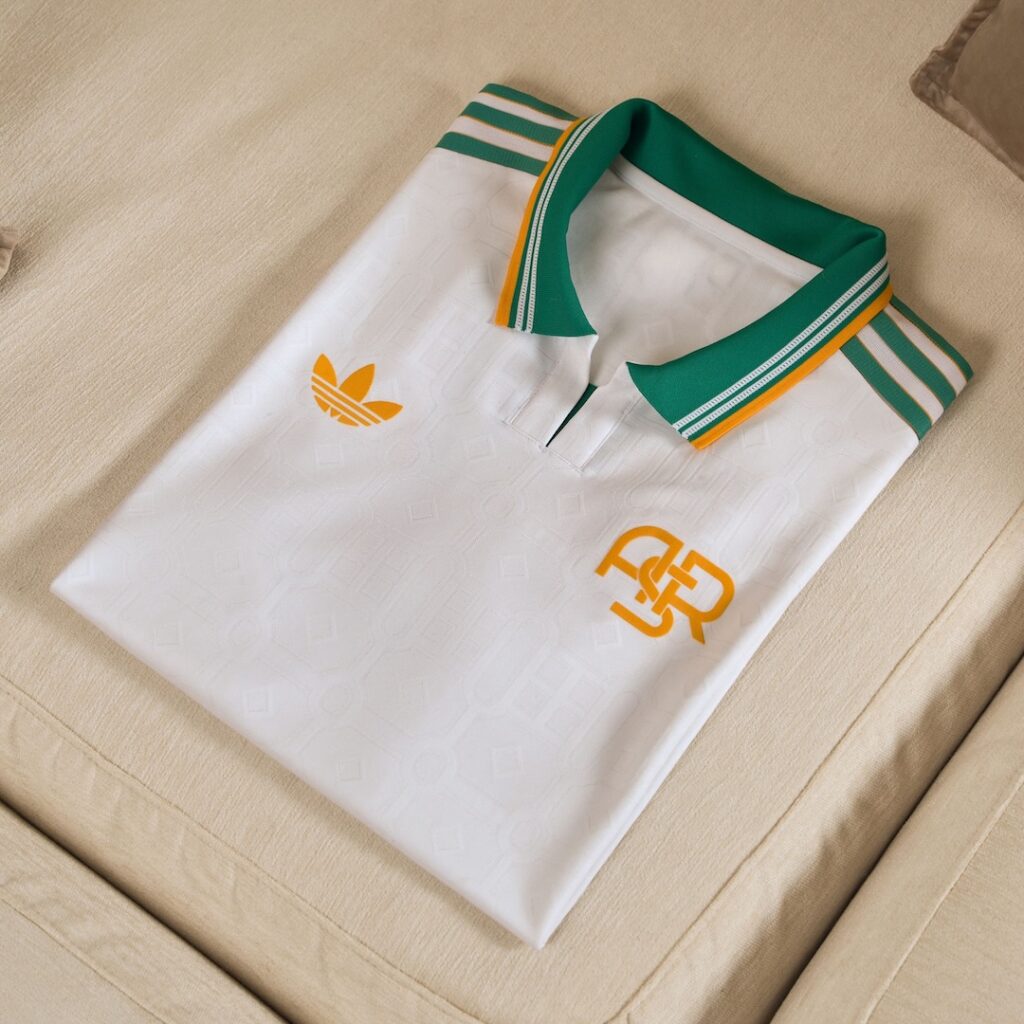 White as Roma jersey with teal and yellow accents folded flat as a square sitting on a cream coloured cushion.