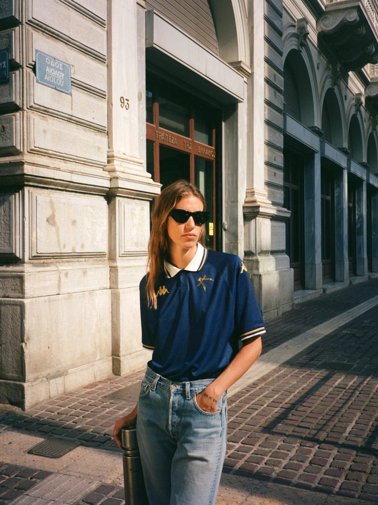 Female model wearing blue Athens kallithea jersey and blue jeans on the streets of Athens. She has one hand in her pocket and is leaning against a street post with the other. She is wearing sunglasses.