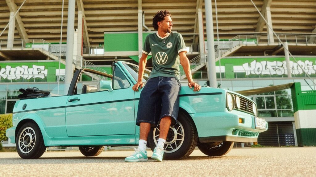 Man wearing jean shorts, turquoise nike sneakers and the green Wolfsburg jersey out front of their stadium, leaning on an old turquoise coloured VW Cabriolet.